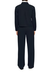 Marineblauwe jas met een kraag en een rugplooien, gecombineerd met een broek met wijde pijpen. Beide stukken hebben een gladde stof en een getailleerd design.