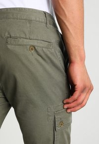 Khaki cargo-byxor med sidfickor och knappdetaljer. Tillverkade av mjukt, hållbart tyg. Har en avslappnad passform och synliga sömdetaljer.