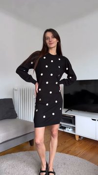 Robe noire à manches longues avec des pois blancs, coupe ajustée, texture douce, longueur au genou, associée à des chaussures noires, posant devant un fond neutre.