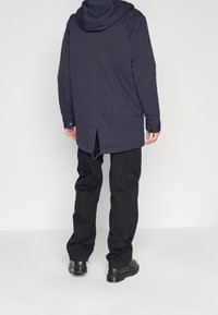 Mies, joka pukeutunut tummaan hupputakkiin, mustiin cargo-housuihin ja mustiin nahkakenkiin, seisoo tasaisen vaalean taustan edessä.