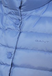 Jasnoniebieska pikowana kurtka puchowa z przeszyciami, okrągłymi guzikami i detalem z logo. Wykonana z gładkiego, błyszczącego materiału o miękkiej fakturze.