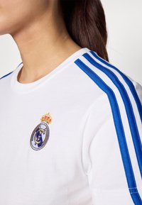 Bílé bavlněné tričko s modrými pruhy na ramenou a vyšitým logem Real Madrid s korunou.