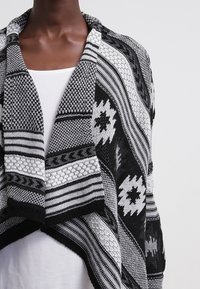 Černobílý šátek s texturovaným úpletem, zdobený geometrickými vzory a splývavým límcem, vrstvený přes bílý top.
