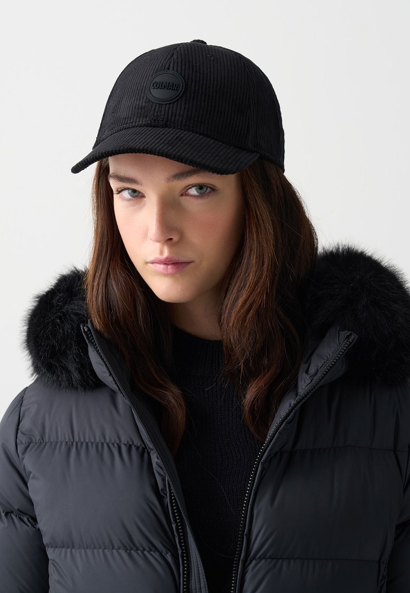 Črna korduro kapa z okroglo črno logotipno nalepko spredaj, nosi jo oseba v bundi s puhastim ovrati.
