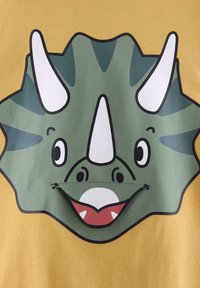 Grøn triceratops ansigt med hvide horn og en smilende rød mund på en sennepsgul stofbaggrund.