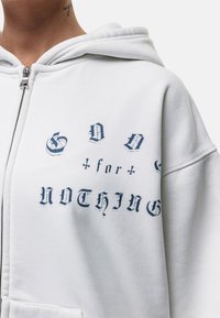 Felpa bianca con zip realizzata in materiale morbido, caratterizzata da testo blu: "DIO + per + NIENTE" in vari font, con una vestibilità comoda e cappuccio.