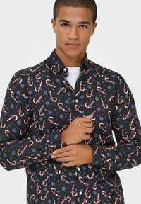 Mees kohandab musta pika varruku särki, millel on punase ja valge piparkoogikinga ning siniste lillede muster, valge alussärgi peal.