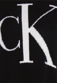 Tissu noir avec de grandes lettres blanches bien visibles. La texture est douce, avec un motif tricoté mettant en valeur une typographie claire et audacieuse.