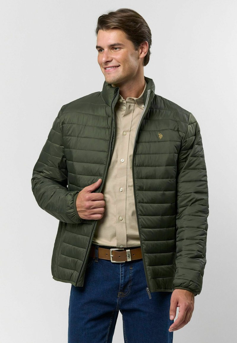 Olivovozelená zateplená bunda s horizontálnym prešívaním, vysokým golierom, predným zipsom a zlatým logom na hrudi. Nosená cez béžovú košeľu.