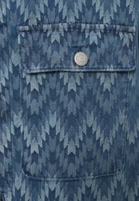 Țesătură din denim albastru cu un model în zigzag, având un buzunar în piept fixat cu un nasture clichet argintiu pentru detaliu.