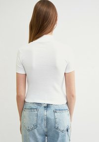 Žena s dlouhými rovnými hnědými vlasy, oblečená v bílém topu s krátkým rukávem a světle modrých džínách s vysokým pasem, stojí zády.