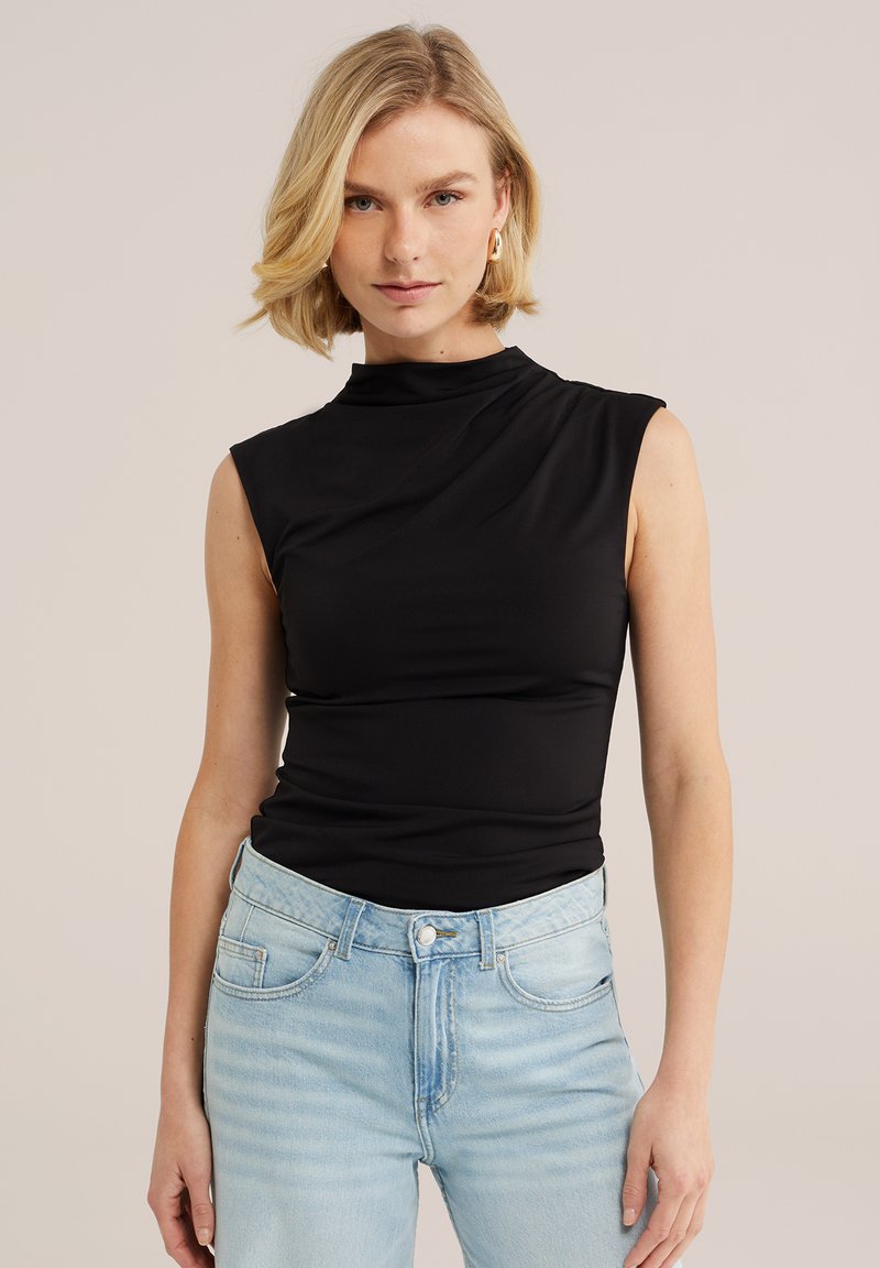 Femme blonde portant un haut noir sans manches à col montant et un jean taille haute bleu clair, debout devant un fond beige.