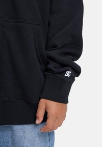 Pravá ruka a dolný rukáv čiernej mikiny s bielym logom DC blízko manžety, v kombinácii so svetlo modrými džínsami.