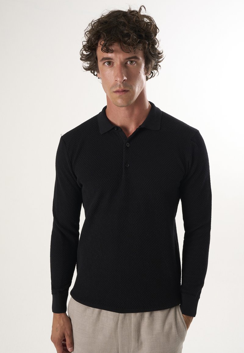 Homme aux cheveux bouclés portant un polo noir texturé à manches longues et un pantalon beige clair, debout devant un fond clair uni.