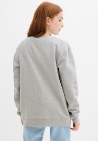 Giovane con capelli rossi che indossa una felpa grigia larga e jeans azzurri, girato leggermente di lato su uno sfondo bianco.
