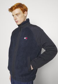 Giacca in pile navy con zip frontale e collo alto, dotata di maniche trapuntate e di un piccolo logo sul lato sinistro del petto. Tessuto morbido e fit rilassato.