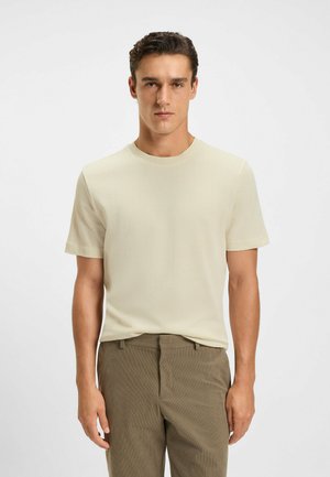 Man wearing cream short-sleeve knit shirt and olive-brown pants standing against a plain white background.