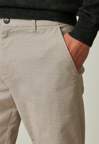 Pantalones beige texturizados con un patrón sutil, que cuentan con un bolsillo frontal y un detalle de botón negro en la cintura.