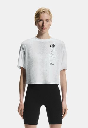 Jeune personne aux cheveux blonds courts portant un T-shirt court blanc avec l'inscription « RUN » et un short de cycliste noir, debout devant un fond uni.