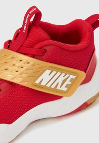 Κόκκινο αθλητικό παπούτσι με επάνω μέρος από αεριζόμενο δίχτυ, χρυσές λεπτομέρειες, μεγάλο λευκό λογότυπο Nike και υφασμένο λουράκι για ασφαλή εφαρμογή.