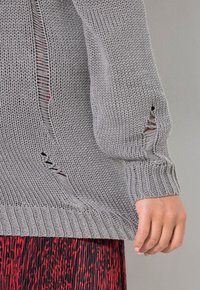 Grauer Strickpullover mit gerippten Bündchen, lockerer Passform und seitlichen Schlitzen. Die Textur ist dick gestrickt, mit subtilen Variationen im Maschenbild.