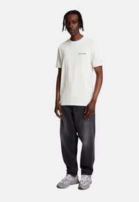 Camiseta blanca de manga corta con texto negro, combinada con jeans sueltos de color gris oscuro y zapatillas grises con un diseño estampado.