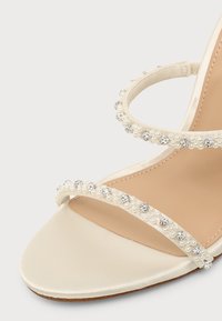 Gros plan d'une sandale beige avec deux brides ornées de petites perles blanches et de strass clairs sur un fond clair.