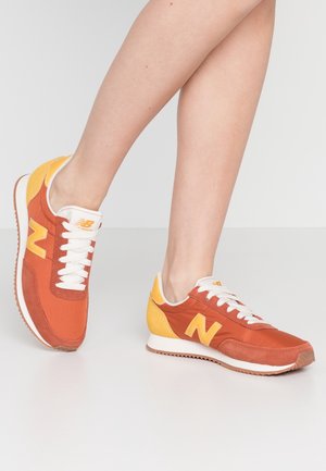 Osoba nosząca pomarańczowo-żółte buty sportowe New Balance z białymi sznurówkami, stojąca na jasnoszarej podłodze.