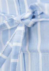 Hellblaues Gewebe mit einem karrierten Muster, das ein weißes Streifenmuster aufweist. Ein gebundener Knoten zeigt die Textur und Weichheit des Materials.