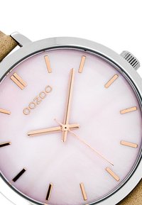 Montre ronde avec un boîtier en argent, un cadran rose clair, des marqueurs et aiguilles en or rose. Un bracelet beige doux complète le design. Esthétique minimaliste.