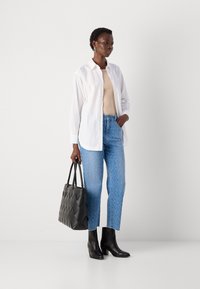 Kvinne med hvit skjorte, beige topp, blå mønstrede jeans, svarte ankelstøvler, og holder en stor svart håndveske foran en hvit bakgrunn.