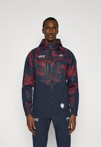 Ανδρικό μπουφάν με κουκούλα σε κόκκινο και ναυτικό μπλε χρώμα, με ελαφρύ σχεδιασμό, τσέπες με φερμουάρ και λογότυπα UFC στο στήθος και το μανίκι.