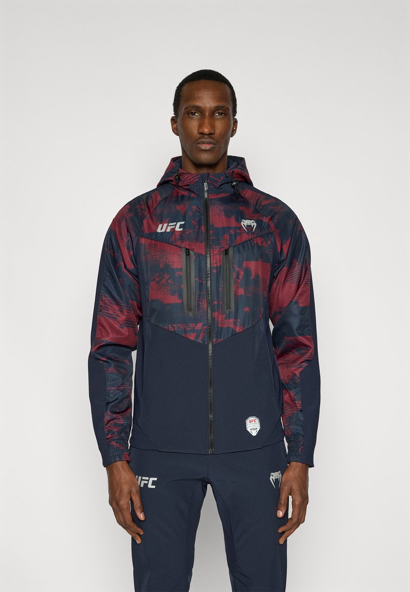 Ανδρικό μπουφάν με κουκούλα σε κόκκινο και ναυτικό μπλε χρώμα, με ελαφρύ σχεδιασμό, τσέπες με φερμουάρ και λογότυπα UFC στο στήθος και το μανίκι.