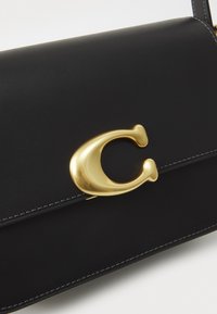 Schwarze Lederhandtasche mit einem auffälligen goldenen Verschluss, klaren Linien und weißen Nähten entlang der Kanten.