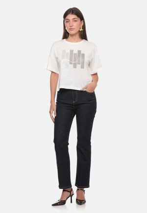 Donna che indossa una T-shirt grafica bianca, jeans scuri, scarpe nere con tacco e grandi orecchini beige, in piedi con una mano in tasca.