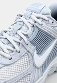 Sapatilha de corrida cinza claro com parte superior em malha e texturizada, detalhes brancos, sistema de atilhos visível e um logótipo proeminente na língua.