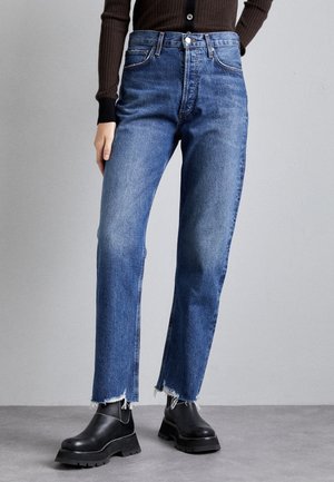 Persona que lleva jeans azul de talle alto con bajos deshilachados, botas negras gruesas y una camiseta marrón ajustada de manga larga sobre un fondo gris.