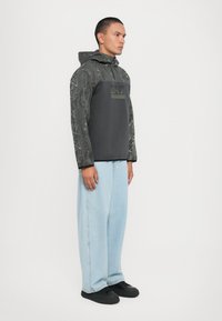 Hættepullover i mørkegrå med mønstrede ærmer. Har et logo på brystet og en afslappet pasform, kombineret med lyseblå jeans og sorte sko.