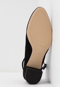 Chaussure à bout pointu en daim noir vue par la semelle beige avec un petit talon noir, posée contre un mur et un sol blancs.