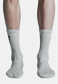 Pilkos ilgos kojinės su raukšlėta tekstūra, papuoštos juodu tekstu šoninėse dalyse, užmautos ant kojų. Audinys atrodo minkštas ir elastingas.