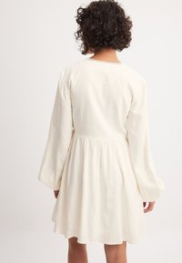 Witte, langmouwige jurk met een gathered taille en een ontspannen pasvorm. De stof is zacht, met een gladde textuur en subtiele details bij de halslijn.