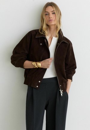 Woman wearing dark brown zip-up jacket, white henley shirt, pinstriped trousers, and a large gold cuff bracelet, standing against white wall.