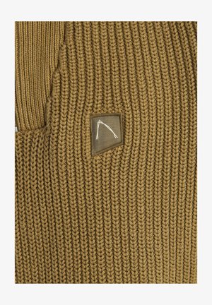 Suéter de punto en un cálido color marrón, con un patrón texturizado. Incluye una etiqueta rectangular con un diseño abstracto en blanco.