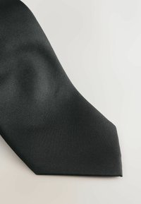 Cravat neagră texturată cu un capăt ascuțit, având un material neted, țesut și un model subtil, pe un fundal deschis.