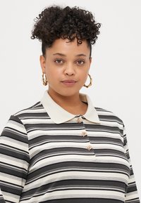 Svītrainā garo piedurknu polo krekls melnā un krēmkrāsā, ar kontrastējošu apkakli, četriem dekoratīviem pogām un mīkstu, teksturētu audumu.