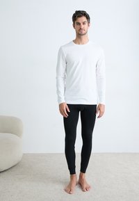 Top branco de mangas longas combinado com leggings pretas justas, design minimalista, textura suave, usado descalço sobre um tapete claro.