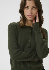 Tmavě zelený pletený cardigan s dlouhými rukávy, kulatým výstřihem a zapínáním na knoflíky. Měkká textura s jemným pleteným vzorem a přiléhavým střihem.