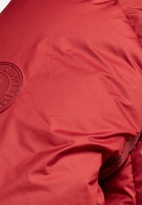 Veste rembourrée rouge avec une texture lisse, comportant un badge circulaire sur l'épaule gauche et une fermeture éclair sur le côté.