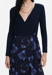 Vestido cruzado de color azul marino con mangas largas; parte superior en textura acanalada, falda con estampado floral oscuro en azules y morados, con detalle de lazo en el costado.