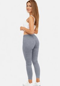 Mujer joven con cabello largo y castaño, que lleva un top corto blanco, pantalones ajustados grises y zapatillas blancas, sonriente y mirando hacia atrás por encima del hombro.